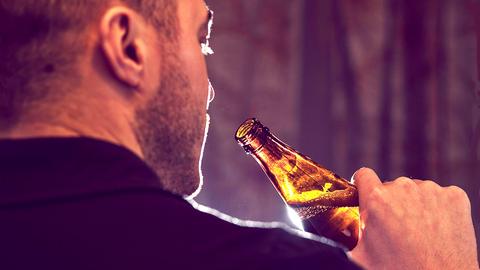 Nahaufnahme eines Mannes von hinten, der eine Flasche Bier zum Mund führt. Licht kommt von hinten, so dass die Flasche beleuchtet ist.