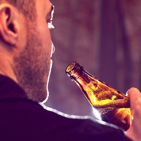 Nahaufnahme eines Mannes von hinten, der eine Flasche Bier zum Mund führt. Licht kommt von hinten, so dass die Flasche beleuchtet ist.