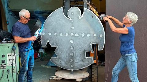 Das Bild zeigt einen Mann und eine Frau, die an einer menschengroßen Engel-Metallskulptur arbeiten