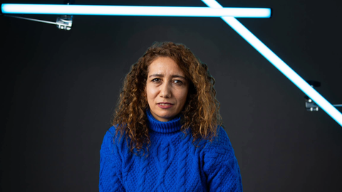 Eine Frau mit braunen Locken sitzt vor einer dunkelgrünen Wand in einem Fotostudio. Sie trägt einen blauen Strickpullover und schaut skeptisch in die Kamera.