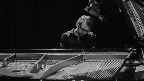 Ein Mann mit Brille spielt auf einem Flügel. Die Innenansicht des Flügels mit den Saiten ist im Vordergrund zu sehen. Der Hintergrund ist dunkel. Der Mann schaut konzentriert nach unten.