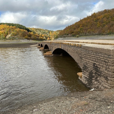 Eine alte Brücke mit vier gemauerten Bögen, unten durch fließt Wasser. Im Hintergrund ist ein Wald zu sehen.