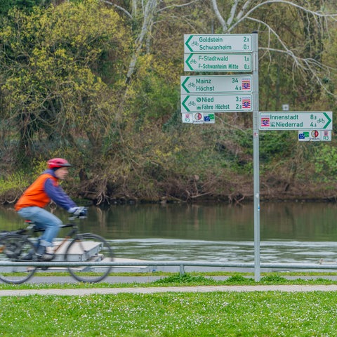 Ein Fahrradfahrer ist unterwegs an einem Fluss. Neben ihm sind Hinweisschilder auf Radwege zu sehen.