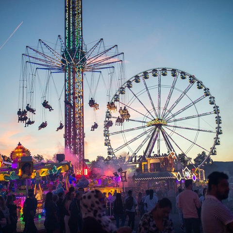 Menschenmenge auf einem großen Platz mit Riesenrad und Karussell vor dem Abendhimmel