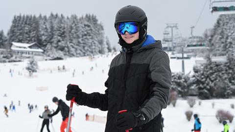 Eine Person in einem schwarzen Skihelm und Skibrille steht auf einer Skipiste. Sie trägt eine schwarze Jacke und hält rote Skistöcke. Im Hintergrund sind weitere Skifahrer, verschneite Bäume und ein Skilift zu sehen. Der Himmel ist bewölkt.