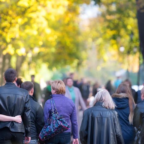 Viele Menschen scharf im Bildvordergrund am unteren Rand, gehend. Im Hintergrund unscharf eine herbstlich gefärbte Baumallee. 