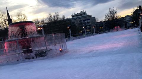 Eine Eisbahn am Wiesbadener Kurhaus mit zerfurchtem Eis und einer runden Aussparung um einen Wasserbrunnen herum in der Mitte. Im Hintergrund ein mehrstöckiges Gebäude.