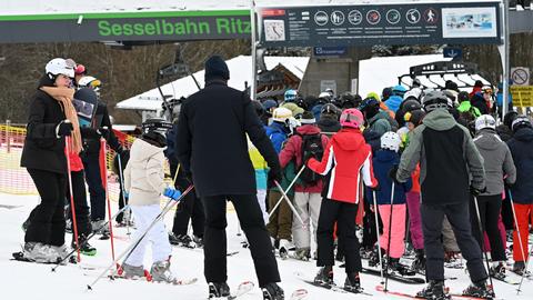 Eine Gruppe von Skifahrern steht in einer Schlange vor der Sesselbahn "Ritzhagen". Die Menschen tragen Skihelme und Winterkleidung. Im Hintergrund ist ein Schild mit Piktogrammen und Uhrzeit zu sehen.