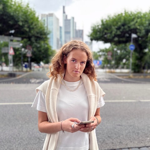 Amelie steht mit Smartphone in der Hand am Sachsenhäuser Mainufer vor der Skyline.