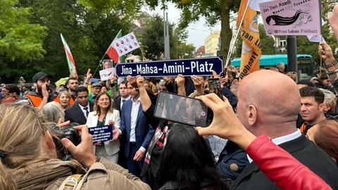 Viele Menschen stehen zusammen. Einer trägt ein Schild mit der Aufschrift: "Jina-Mahsa-Amini-Platz".