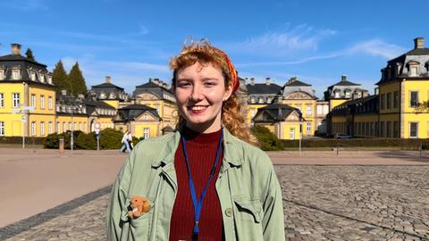 Eine junge Frau mit buntem Kopftuch, rotem Pullover und einem Teddybären in der Brusttasche ihrer Jacke lächelt vor dem Schloss in Bad Arolsen in die Kamera