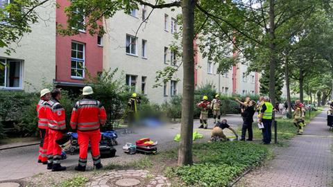 Das Bild zeigt einen breiten Bürgersteig zwischen mehreren hohen Wohnhäusern und einer Reihe von Bäumen. Auf dem Bürgersteig sind Feuerwehrleute im Einsatz und Rettungskräfte - offenbar hat es in dem Haus dahinter gebrannt.