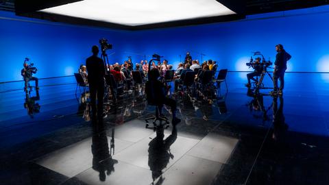 Ein beleuchtetes Studio, viele Menschen sitzen im Kreis unter einem weißen Licht, Kameras drumherum