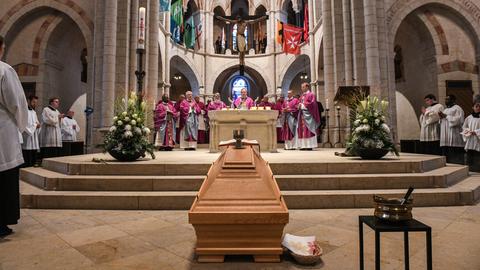 Das Bild zeigt den Limburger Dom während des Requiems für Altbischof Franz Kamphausen. Im Vordergrund ist ein Sarg aus hellem Holz zu sehen, darauf steht ein Messing-Krug. Am Altar steht der derzeitige Bischof Georg Bätzing und predigt. Er ist umgeben von weiteren kirchlichen Würdenträgern, sie alle tragen Gewänder Weiß und Lila. Im Hintergrund ist eine Skulptur von Jesus am Kreuz zu sehen.