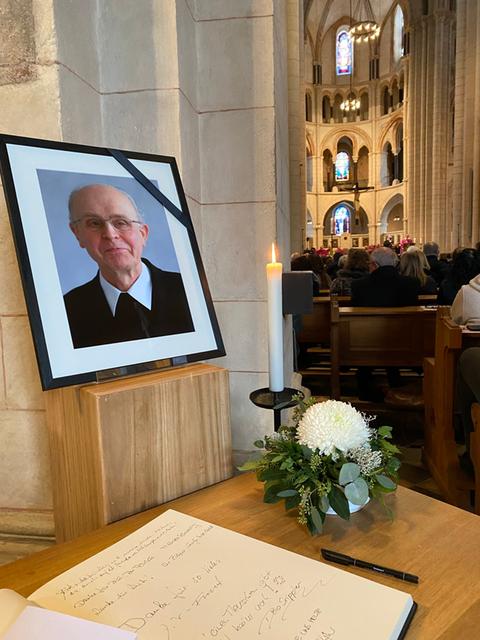 Das Bild zeigt den Limburger Dom während der Beisetzung des früheren Bischofs Kamphaus. Zu sehen ist ein schwarz gerahmtes Bild eines alten Mannes mit Brille, wenig Haaren und dunklem Pullover mit hervorschauendem hellem Hemdkragen. Über der rechten Ecke des Bildes ist ein schwarzes Band gespannt. Auf einem Holztisch davor befinden sich ein Blumengesteck mit weißen Blüten sowie ein Gästebuch, in dem sich Besucher handschriftliich eingetragen haben.