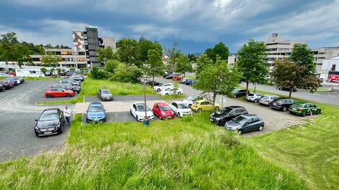 Blick auf einen Parkplatz mit vielen Autos, Gebäude und kleinerer Grünflächen aus einer halben Vogelperspektive.