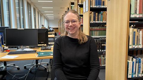 Eine Frau Mitte 40 mit blonden Haaren und einer Brille sitzt zwischen Bücherregalen in einer Uni-Bibliothek