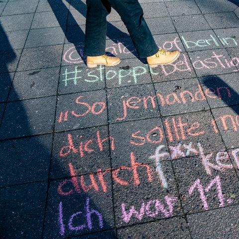 Bunte Kreideaufschrift auf einer Straße. Sie lautet: "Jemanden wie dich sollte man mal durchf**cken. Ich war 11." Dahinter angeschnitten die Füße einer Person, die daneben steht.