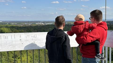 Zwei Männer und ein Kind auf dem Arm eines Mannes (von hinten zu sehen) stehen auf einer Aussichtsplattform und schauen in die Ferne. Im Hintergrund weite Landschaft und blauer Himmel mit Wolken.