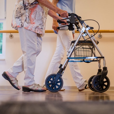 Im Vordergrund schiebt eine ältere Frau ihren Rollator durch einen Pflegeheim-Korridor. Im Hintergrund ist zu erkennen, dass sie von einer Pflegerin in einem blauen Kittel begleitet wird. 