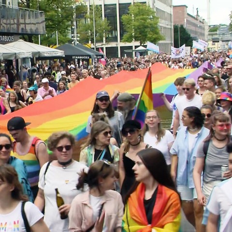 Teilnehmerinnen und Teilnehmer an der CSD-Parade in Darmstadt gehen durch eine Straße mit einer riesigen Regenbogenflagge