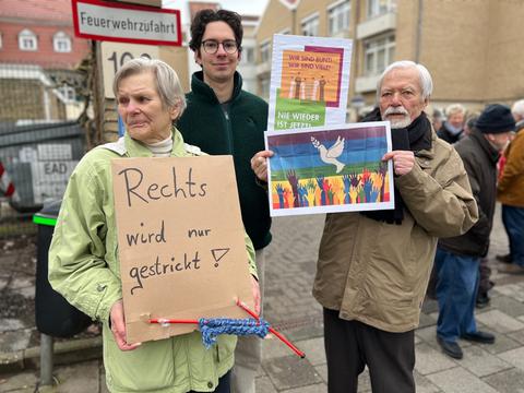 Frau mit Plakat mit Stricknadeln und Aufschrift "Rechts wird nur gestrickt", daneben ewin junger Mann und ein älterer Herr