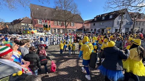 Eine große Menschenmenge nimmt an einer Karnevalsveranstaltung teil. Im Vordergrund sind viele Personen in gelben Kostümen zu sehen, einige tragen gelbe Hüte. Im Hintergrund ist ein großer Festwagen mit einem Krönchen und darauf sitzenden Personen, umgeben von Menschen in blau-weißen Uniformen. Im Hintergrund sind Häuser. Links oben sind Fahnen zu sehen. Rechts an einem Gebäude ist das Geschäftsschild "Euronics" erkennbar.