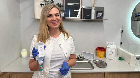 Eine Frau mit blonden Haaren lächelt in die Camera. Sie trägt einen Arztkittel, blue Gloves und hält einen Infusionbeutel in der Hand.