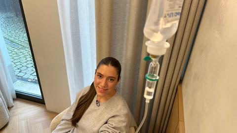 Eine junge Frau mit dunklen Haaren lächelt. Das Bild ist in der Vogelperspektive fotografiert und man sieht einen Infusionsbeutel, der von oben der Frau an den Arm gelegt ist.