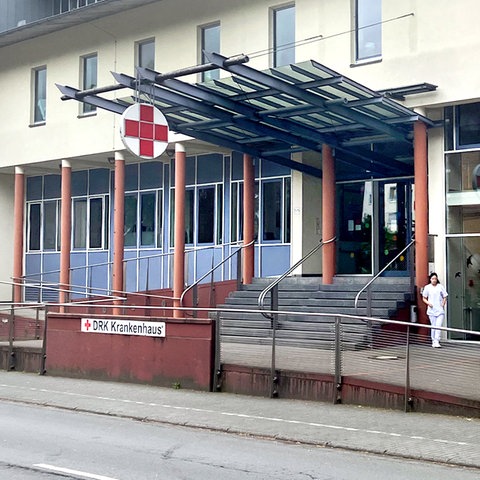 Krankenhausgebäude von außen. Vor der Treppe steht ein Schild "DRK Krankenhaus". Über dem Eingang ein großes rotes Kreuz auf weißem runden Grund