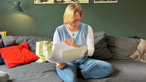 Lee-Ceshia Lemcke sitzt auf dem Sofa in ihrer Wohnung und blättert durch Arztunterlagen. Sie hat blonde, kinnlange Haare und trägt eine Brille.