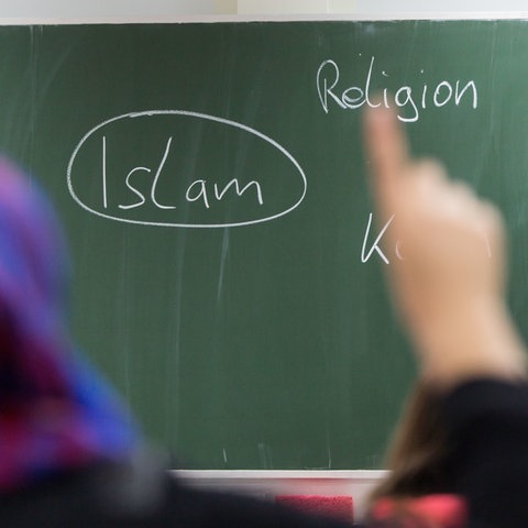 Eine Schülerin mit Kopftuch meldet sich im Unterricht. An der Tafel prangt das Wort "Islam"