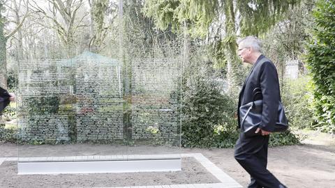 Ein Mann im dunklen Anzug und  mit Tasche unter dem linken Arm geht auf die transparente Stele mit den Namen der Opfer zu.