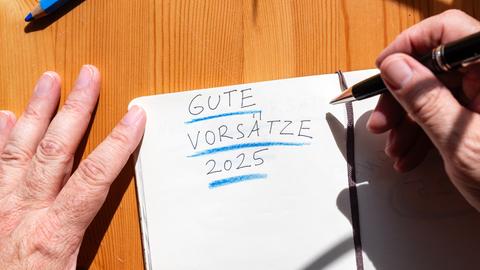 Ein Heft auf einem Tisch, auf dessen aufgeschlagener Seite "Gute Vorsätze 2025" geschrieben steht. Umrahmt wird das Heft von zwei Händen. 