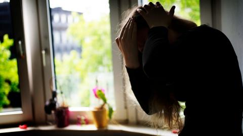 Symbolbild: Eine Frau sitzt an einem Fenster in einer Wohnung und hält sich schützend die Arme vor das Gesicht.