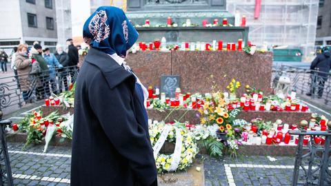 Eine Frau steht im Vordergrund vor den vielen Trauerbekundungen in Form von Blumen, Kränzen und Kerzen an einem Denkmal in Hanau. Im Hintergund sind weitere Menschen zu sehen.