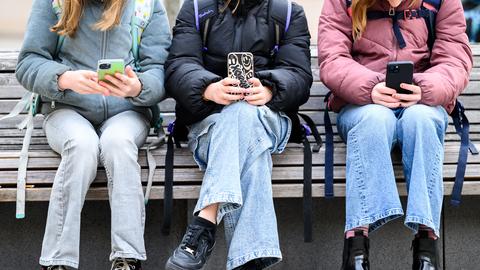 Drei Kinder mit Schulrucksäcken sitzen auf einer Holbank und haben jeder ein Handy in der Hand.