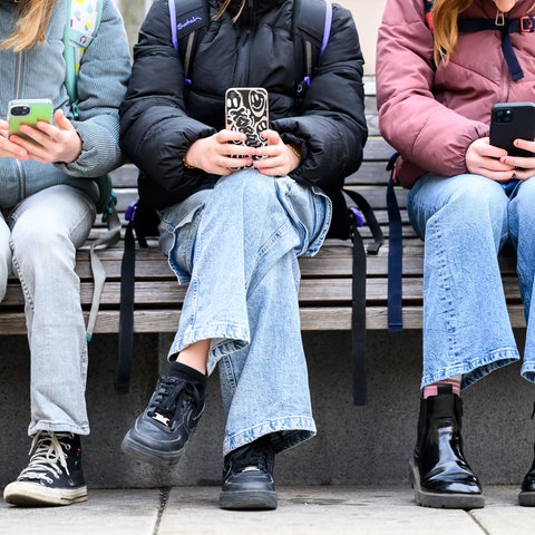 Drei Kinder mit Schulrucksäcken sitzen auf einer Holbank und haben jeder ein Handy in der Hand.