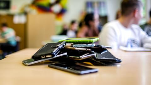 Viele Smartphones (im Bildvordergrund scharf) liegen auf einem Stapel auf einem Tisch in einem Klassenraum (im Hintergund unscharf).