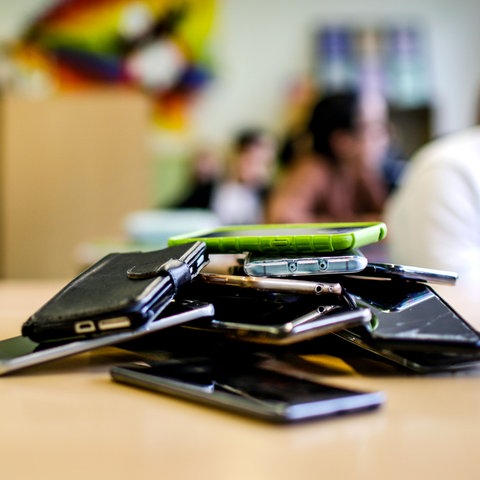 Viele Smartphones (im Bildvordergrund scharf) liegen auf einem Stapel auf einem Tisch in einem Klassenraum (im Hintergund unscharf).