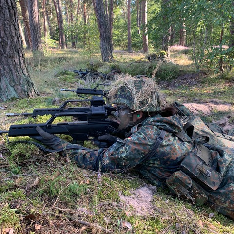 Eine Rekrutin in Tarnuniform liegt auf dem Waldboden und zielt mit einem Gewehr