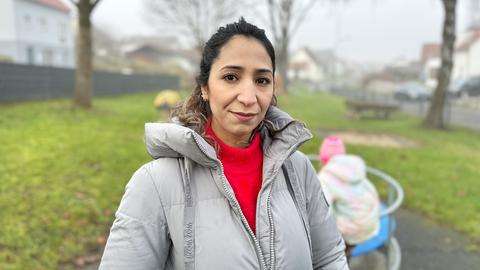 Eine Frau in grauer Jacke steht auf einem Spielplatz (im Hintergrund unscharf spielende Kinder auf einem Karussell) und blickt ernst in die Kamera.