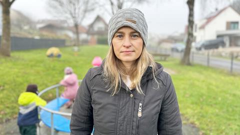 Eine Frau in grauer Jacke und grauer Mütze steht auf einem Spielplatz (im Hintergrund unscharf spielende Kinder auf einem Karussell) und blickt ernst in die Kamera.