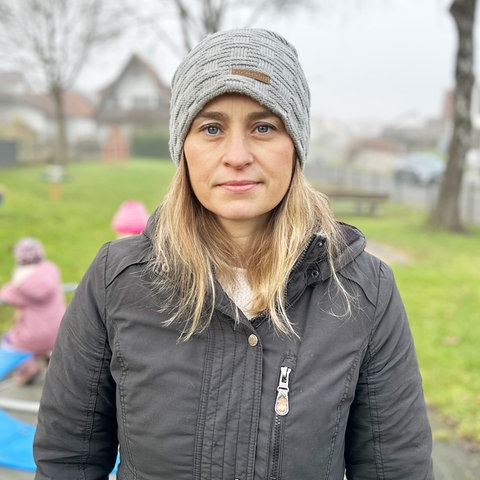 Eine Frau in grauer Jacke und grauer Mütze steht auf einem Spielplatz (im Hintergrund unscharf spielende Kinder auf einem Karussell) und blickt ernst in die Kamera.