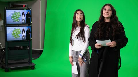 Zwei junge Frauen stehen in einem Fernsehstudio vor einer grünen Wand.