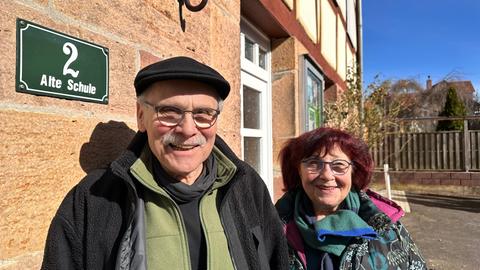 Ein Mann mit Hut und eine Frau mit bunter Jacke stehen vor einem alten Haus. Neben ihnen ist ein Schild mit "Alte Schule" zu sehen.