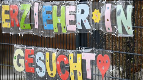 An einem Zaun hängen bunte Buchstaben, die die Wörter "Erzieher*in gesucht" ergeben. 