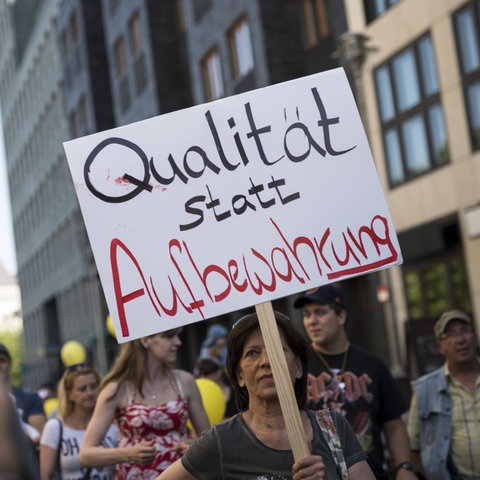 Erwachsene und Kinder in einer Straße beim Demonstrieren für bessere Kinderbetreuung. Jemand hält ein Schild hoch mit der Aufschrift "Qualität statt Aufbewahrung".