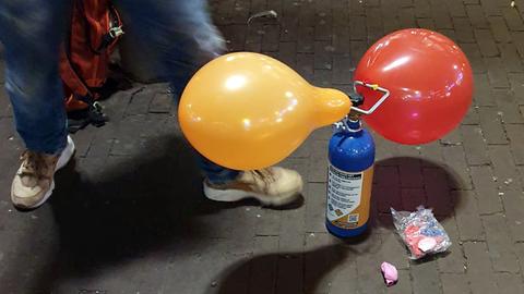 Auf dem Boden steht eine Gaskartusche, an der ein orangfarbener und ein roter Luftballon hängen. Daneben sind die Beine eines Menschen zu sehen, der danebensteht.