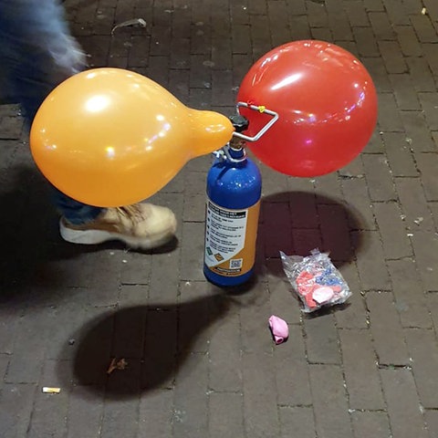 Auf dem Boden steht eine Gaskartusche, an der ein orangfarbener und ein roter Luftballon hängen. Daneben sind die Beine eines Menschen zu sehen, der danebensteht.